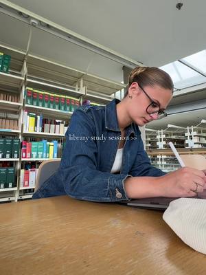 A young woman wearing glasses and a denim jacket sits studying at a desk in a library. She's writing on a tablet and a macbook is on the desk. Bookshelves fill the background.