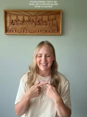 A woman with long blonde hair smiles and looks towards the camera. She is wearing a light colored t-shirt. She is pointing her index fingers to each other in front of her chest. Behind her is a light green wall with a wooden carving mounted to it. The carving depicts a scene of people seated around a long table.