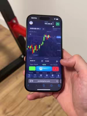 A person's hand holds a black smartphone displaying a trading app. The app’s interface shows a candlestick chart and trading options. The background is a cream-colored wall and a glimpse of a red object, perhaps a piece of exercise equipment.