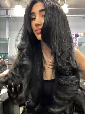 A woman with long, dark, wavy hair poses for a selfie in what appears to be a hair salon. Her hair frames her face, and she's wearing a black top. The background shows mirrors and salon equipment, and there are bright overhead lights. The overall composition suggests a focus on her new hairstyle.