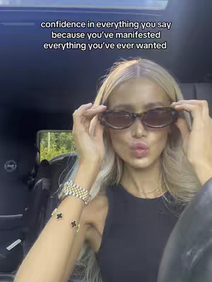 A woman with blonde hair and sunglasses poses for a selfie while seated in a car. Text overlayed on the image gives a motivational message. The overall composition uses natural light, the top of the car can be seen.