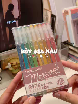 The image shows a close-up of a person's hands holding a clear plastic case containing a set of colorful gel pens. The pens are arranged vertically, offering a spectrum of colors from blue to red. Below the pens, there is a brown label with the brand name "Morandi" in a stylized script, with text in Chinese. The background is out of focus but suggests a desk or table setting with other stationary and possibly a computer screen displaying a clock at 19:00.