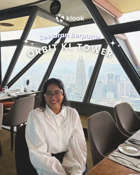 A woman with dark hair and glasses sits in a restaurant with large windows overlooking a city. She is smiling at the camera. The restaurant has a modern design with dark wood and neutral-toned chairs. The city skyline is visible in the background.
