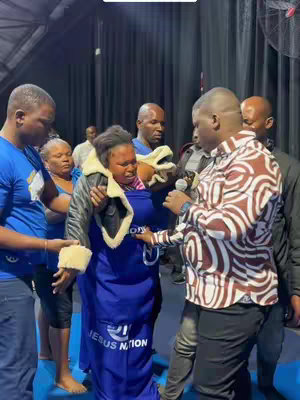 A group of people are gathered in what appears to be a church or event space. One person is speaking into a microphone, while others support another person. The main subject is a woman in a blue dress with a blue cloth draped over her shoulders, surrounded by others. The color palette is muted with a mix of skin tones, blue, brown, and beige.