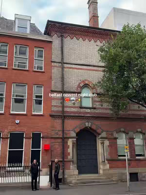 The cover image shows a street view of a building facade in Belfast. The building has red brick and stone construction. A black door is centered at the bottom, with two individuals standing to the sides. The sky is overcast. The text overlay reads, 'belfast newest night club' with graphic elements.
