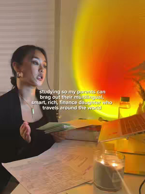 An Asian woman is seated, facing slightly to the left. She has dark hair, and is wearing a black blazer and a necklace. She is in front of a desk with papers and a laptop, as well as a lit candle. A bright orange and yellow light is projected behind her, possibly from a sunset lamp. The text overlayed says: studying so my parents can brag out their multilingual, smart, rich, finance daughter who travels around the world.