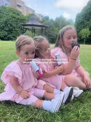 Three young girls are seated on green grass, with a small gazebo visible in the background. They are all wearing similar pink dresses. One girl holds a microphone to the left, and the other is eating from a black container. Text is overlaid on the image.