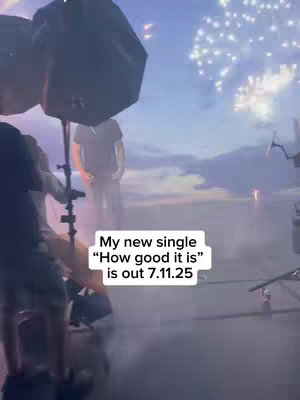 The image shows a behind-the-scenes look at a music video shoot. It features a man standing in the background, partially obscured by lighting equipment. The setting appears to be a studio with a projected nighttime scene, complete with fireworks. A person is visible, with the text overlayed at the bottom. The composition blends backstage elements with a cinematic backdrop.