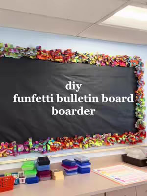 A classroom scene featuring a bulletin board with a black background and colorful, confetti-like border. Below the board, a counter holds trays, containers, and printed papers.