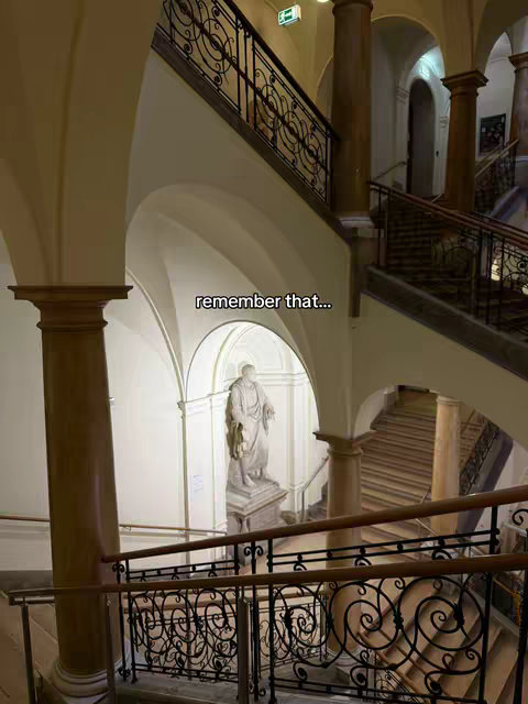 An interior shot featuring an ornate staircase with a statue under a dome. The image has a warm, inviting color palette with beige walls, brown woodwork, and golden lighting. The composition is symmetrical, drawing the eye from the foreground staircase up to the arched ceiling and the statue. The railings have scrollwork, adding to the classical feel.