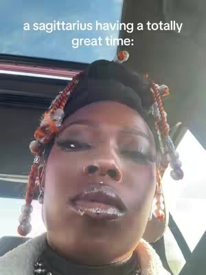 A close-up selfie shows a woman in a car. The woman is looking up and to the side, with a slightly open mouth. She has multiple braids with decorations and is wearing multiple accessories and makeup.