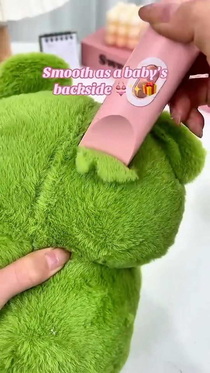 The image shows a close-up of a fluffy, bright green fabric, with a pink grooming tool seemingly shaving or trimming it. The tool is held by a hand, and text overlayed reads 'Smooth as a baby's backside'. The background appears to be a clean, light-colored surface.