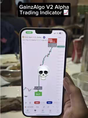 The cover image showcases a smartphone displaying a trading application interface. The screen is dominated by a colorful line graph indicating market fluctuations, with 'SELL' and 'BUY' buttons visible. A 3D rendered skull icon is centrally placed, apparently indicating market trend or risk. Text overlays at the top reads 'GainzAlgo V2 Alpha Trading Indicator'. The background appears to be indoors, as indicated by the blurred features. The phone is held in someone's hand.
