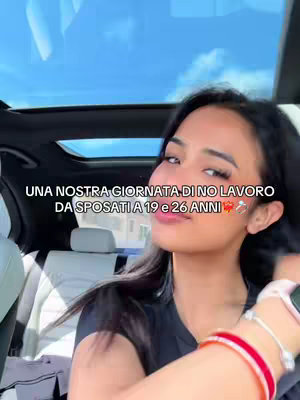 A woman is seated inside a car, likely a front seat, looking towards the camera. She has long dark hair and is wearing a black top and a watch. The background shows a sunny sky through the car's sunroof, and text overlays at the bottom of the image.
