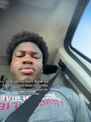 A selfie shot of a young Black man inside a vehicle, angled from below. The man has short, curly hair and a somewhat pensive expression with his lips pursed. He is wearing a gray shirt with a logo on it and a seat belt. Text is overlaid on the image.