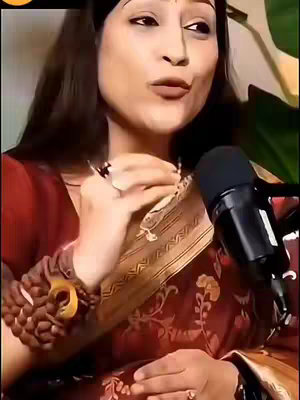 A woman in a red and gold patterned saree is holding her hand up, appearing to be reading it. She has dark hair, brown eyes, and is wearing bracelets and a ring. A microphone is visible, suggesting she's speaking or recording. The background is blurred, with hints of a green plant.