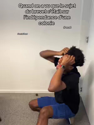 A young Black man is crouched on a low surface, with his hands on his head in a gesture of distress. Above him, text overlays read in French. The background is a plain white wall.