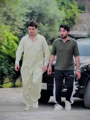 Two men walking side by side outdoors. The man on the left is wearing a light-colored outfit; the man on the right is wearing a green shirt and black pants.