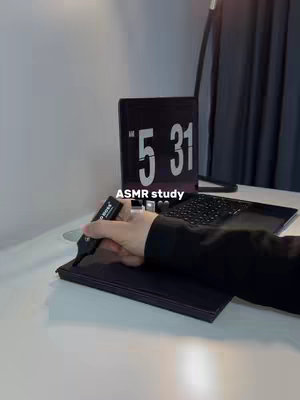 A close-up shot of a desk with a laptop, clock and a person's arm holding a pen over a notebook. The laptop screen displays a clock that reads '5 31'. The image is taken in a dark aesthetic style.