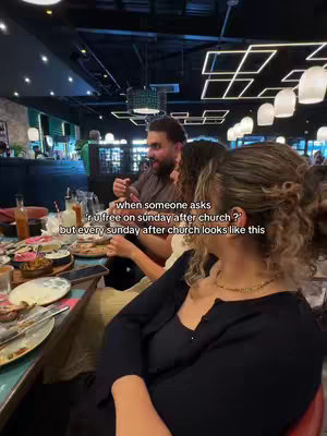 A photo of people at a table in a restaurant. The focus is on three people, with others blurred in the background. A man wearing a brown shirt is speaking animatedly. The woman next to him has her back to the camera and is wearing a black cardigan. The setting has warm lighting, and the table is laden with food and drinks, suggesting a meal. Text is overlaid on the image.