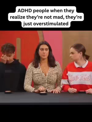 The image is a vertical video frame featuring three people seated behind a black table. At the top, there is a white banner of text. The background is a visually noisy mix of red and pink walls, adding intensity. The subjects, from left to right, are a young man, a woman in the middle, and a young woman on the right. 

The young man on the left is wearing a black hoodie, and his hand is covering his eye. In the center is a woman wearing a printed blouse, gazing solemnly forward. The woman on the right wears a red and white striped shirt, with a contemplative look. Each person has a small black item in front of them.