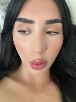 A close-up shot of a woman's face, seen from a slightly downward angle. Her dark hair frames her face. She has full, glossy lips and prominent eyebrows. The focus is on her face, with soft lighting highlighting her features. The background is blurred, suggesting an intimate setting.