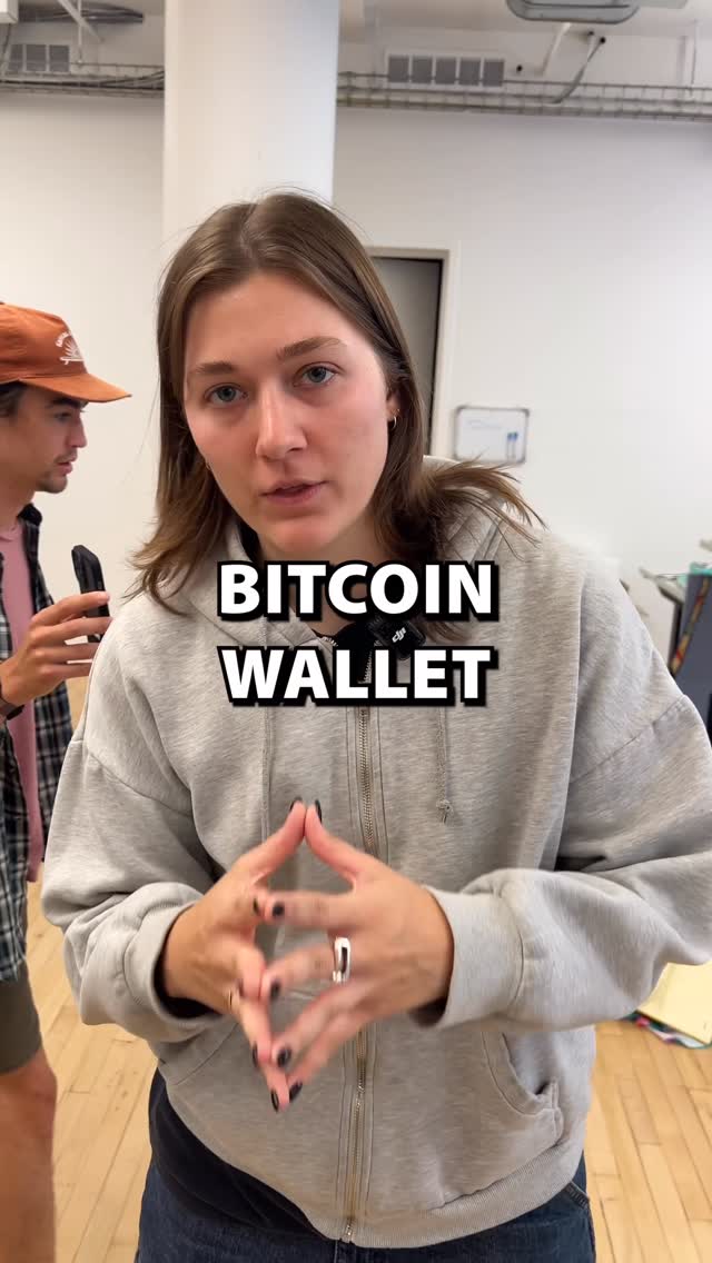 The cover image shows a woman standing indoors, likely in an office setting. She's wearing a gray hoodie and her hands are clasped together, fingers intertwined, as if she's making a point. Text overlayed on the image reads 'BITCOIN WALLET' in bold, white letters. A man is partially visible in the background, wearing a baseball cap, and a microphone is attached to the woman's shirt. The overall composition indicates a video about Bitcoin.