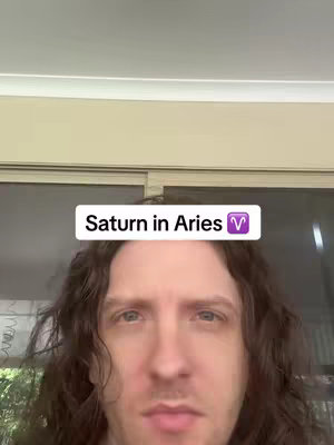 A close-up shot of a man with long, wavy brown hair. He is looking directly at the camera with a neutral expression. Above his head is a rectangular white box with the text 'Saturn in Aries' and a purple Aries symbol, set against a background of a neutral color.