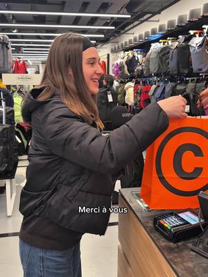 A young woman, smiling and giving a bag to the camera, stands at a shop counter. The background shows shelves of merchandise in retail lighting. On the bottom of the frame is a text box with the message 'Merci à vous'.
