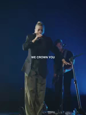 A person, likely a performer, stands on a stage and holds a microphone while singing. The background is dark with a bluish hue, and another person is visible playing an instrument. White text overlays the image.