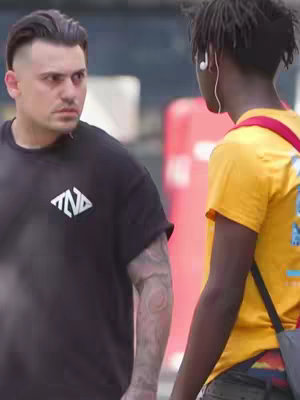The cover image shows two men facing each other outdoors. The man on the left, in a black t-shirt with a logo, has a serious expression. The other, wearing a yellow shirt and earbuds, faces the first man. The background is blurred.