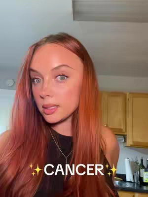 Close-up of a young woman with red hair, gazing towards the camera. The background includes kitchen elements like cabinets. The text overlay at the bottom says 'CANCER' with star embellishments.