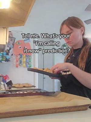 A young woman with auburn hair, wearing a black t-shirt, is in a kitchen setting. She is holding a baking tray with cookies, appearing to take them out of the oven. The background includes a kitchen counter, appliances, and a decorative piece on the refrigerator. Text overlayed on the image says 'Tell me. What's your I'm calling it now' prediction?'