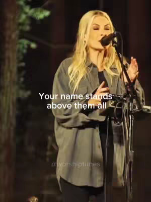 A woman with blonde hair is singing into a microphone on a stage. The lighting is dim, and the background is blurred. Text overlayed on the image reads "Your name stands above them all."