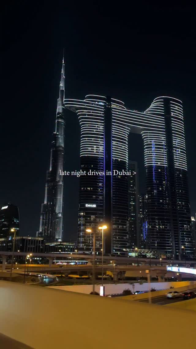 A nighttime cityscape of Dubai featuring illuminated skyscrapers. The Burj Khalifa stands tall on the left side, with two other buildings with distinctive arch-like structures taking a central position. Roads are visible, with streetlights adding to the nighttime ambiance.