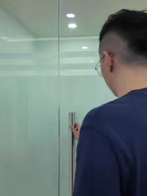 A person is shown from the back, wearing a navy blue shirt and glasses, reaching toward a glass door handle. The scene is set in an office or similar environment, with a clean, minimalist aesthetic. The camera angle is focused, showing the interaction with the door.