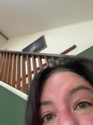 A close-up shot captures a woman's face, with her eyes and top of her forehead dominating the frame. Parts of a staircase and an artwork on the wall are partially visible in the background.