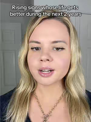 A woman with blonde hair and makeup is centered in the shot, framed from the chest up. Above her head is overlaid text, while she looks towards the camera with a slightly open mouth. She is wearing a necklace and a dark top, with a neutral background that fills the edges.