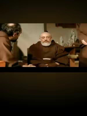 The cover image shows a scene from a film. In the foreground, a man in a brown habit is seated, facing slightly to the right. His head is balding, with white hair around the back and sides, and a white beard. He has a serious expression. Two other men in similar brown habits are partially visible, one on the left and one on the right, suggesting a setting with other figures. The overall lighting is dim, creating a subdued atmosphere, and the background is blurred.