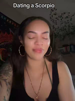 A woman with dark hair and hoop earrings is centered in the frame. The background is blurred but includes a patterned tapestry and some plants. Text overlayed at the top says, "Dating a Scorpio."