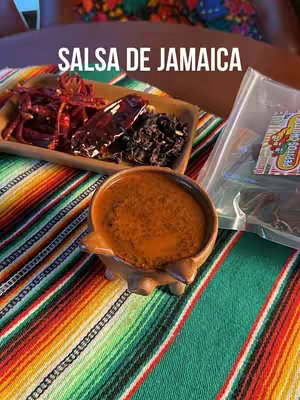 The cover image shows a close-up shot featuring a small, brown, handcrafted bowl filled with a brownish-red sauce set on a colorful, patterned fabric. To the left of the bowl, there is a wooden tray displaying dried red chili peppers and spices. To the right, a transparent plastic bag is partially visible, containing more dried ingredients. The background is slightly blurred, but it seems to be a wooden surface in a room. A large text overlay at the top of the image says 'SALSA DE JAMAICA'. The overall composition suggests a culinary theme, possibly related to making salsa, with a focus on ingredients and presentation.