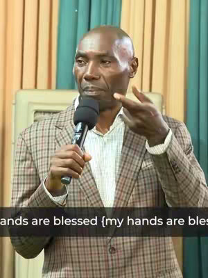 A man in a plaid blazer holds a microphone, gesturing with his right hand as he speaks. The background is a neutral color with vertical striped curtains. Text overlaid towards the bottom reads "hands are blessed".