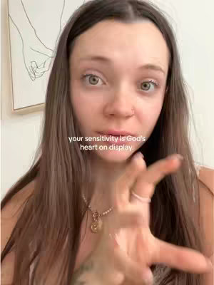 The image shows a close-up of a young woman with long brown hair, a nose piercing, and a necklace. She is looking directly at the camera with a slightly surprised expression. She is gesturing with her right hand, as if emphasizing a point. The background includes a blurred image of a hand-drawn piece of art in a white frame.