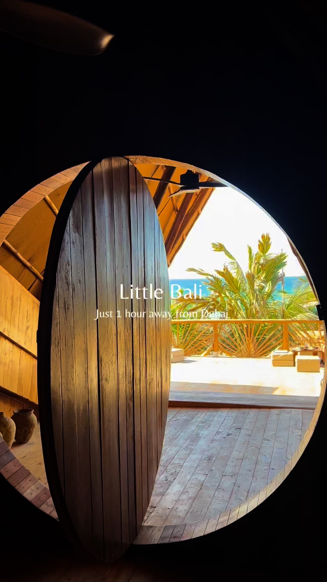 The image is framed through a wooden archway, resembling a stylized boat shape. Inside, a wooden deck leads towards an open view of lush green foliage and a serene sea under a bright sky. The archway is dark and contrasted against the bright exterior, with the wooden door slightly ajar.