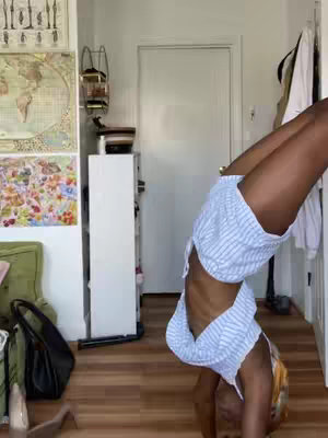 A person is doing a handstand indoors, with their feet against a wall. They are wearing light blue striped shorts and a white top. The background includes a door, a dresser, a wall with artwork, and miscellaneous items.