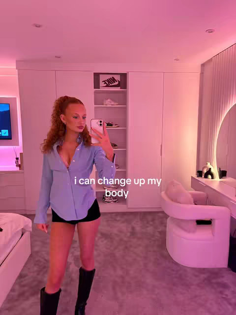 A young woman with auburn hair standing in a pink-lit room. She is facing the camera and holding a phone up to capture a selfie. In the background, there's a white closet, and shoes on the shelves, the overall image has a clean, modern aesthetic.