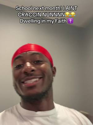 A selfie-style shot featuring a young Black man smiling widely with a red durag on his head. The background is a plain light wall. Text overlayed in the image says, School next month! I AINT CRACCIN NUNNNN 😂😭 Dwelling in my Faith ✝️.