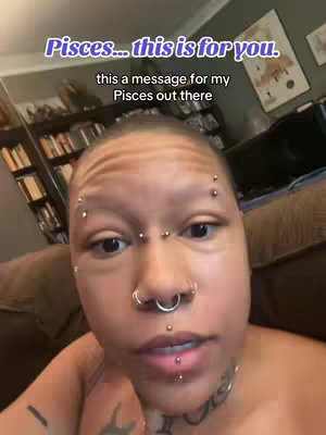 A close-up shot of a person looking at the camera. They have numerous facial piercings, including eyebrow and lip piercings. A tattoo is visible on their neck. The background is blurred, suggesting an indoor setting.