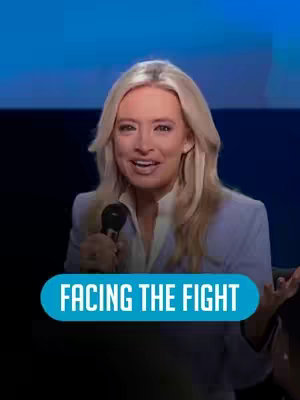 A well-lit medium shot features a woman with long, blonde hair and a light-colored blazer, speaking into a microphone. A teal banner labeled "FACING THE FIGHT" is prominently displayed below her. The background is blurred, with a gradient of blues giving the impression of a formal stage setting.