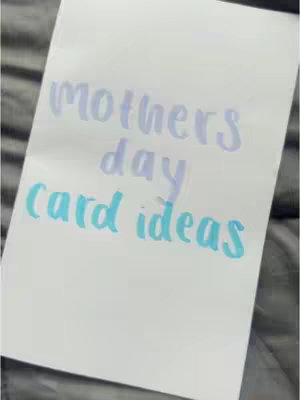 A close-up shot of a white sheet of paper angled towards the top right corner, resting on a gray surface with blurred fabric details. Handwritten in the center in a large, friendly handwritten font are the words "mothers day card ideas". The word "mothers" is in light purple, "day" is in a slightly darker purple, and "card ideas" is in a turquoise blue, each word stacked vertically.